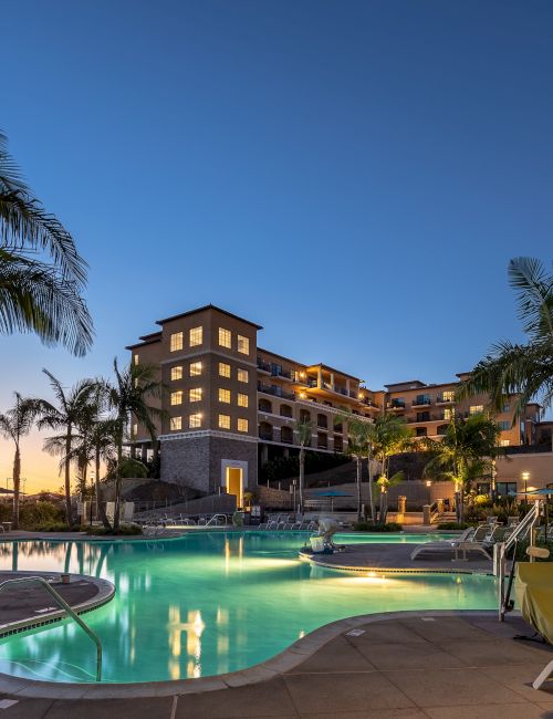 A resort pool area at dusk with palm trees, lounge chairs, and a multi-story hotel building glowing in warm lights; tranquil evening vibes.