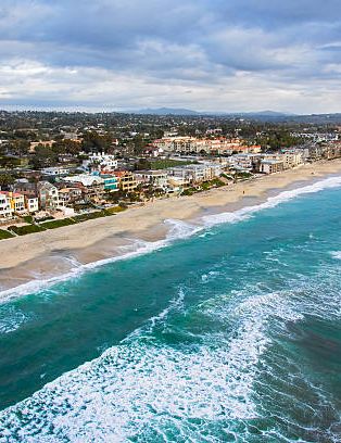 Aerial view of a sunny coastal town with colorful houses along a sandy beach; turquoise waves crash onto the shore, horizon stretches far.