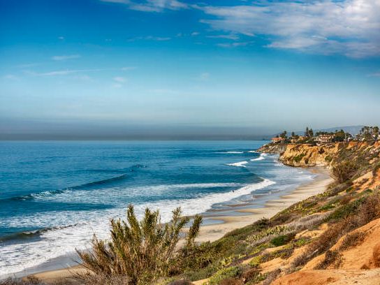 A coastal cliffside with a blue ocean, gentle waves, sandy beach, and some low vegetation along the rocky shoreline, under a clear sky.