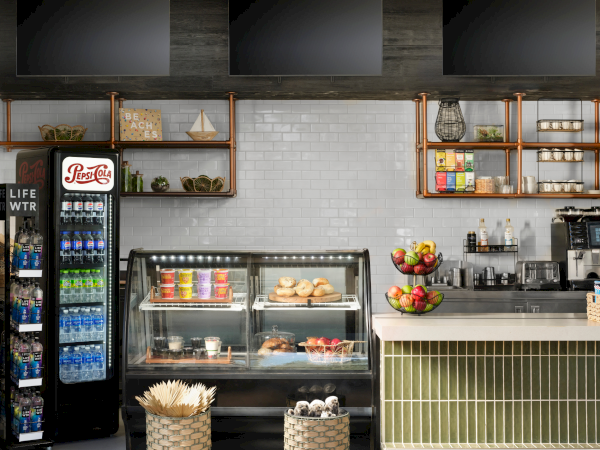 A modern cafe interior with a display case of pastries, baskets, fruit bowls, a beverage fridge, and open wooden shelves above the counter.