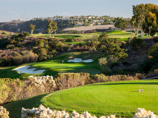 A scenic golf course perched among rolling hills, with green fairways, sand bunkers, and trees, under a clear sky.