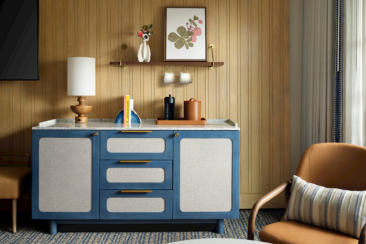 A retro blue cabinet with gray panels sits against a wood paneled wall, topped by a lamp, books, and small decor; a brown chair and window nearby.