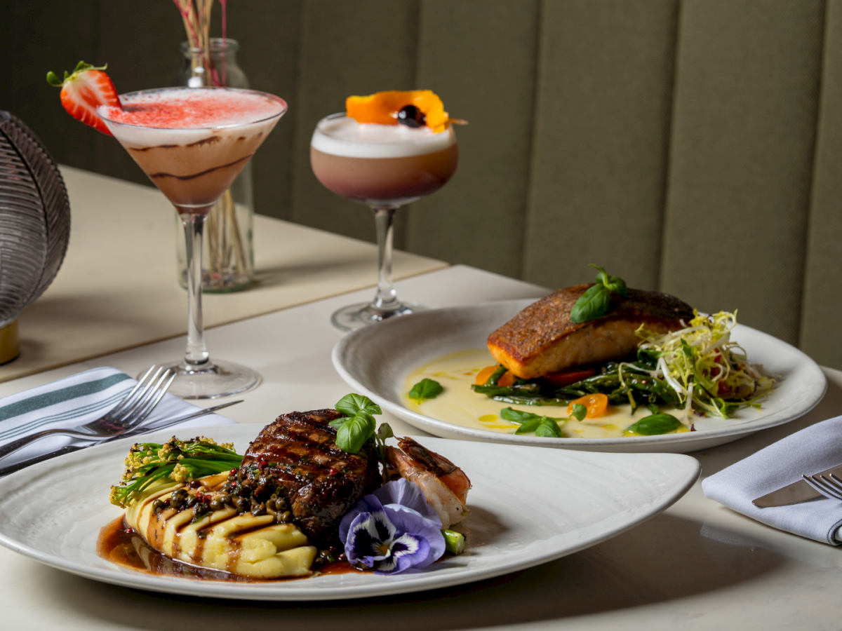 Two fancy dishes with steak and fish, plus two cocktails on a chic table&mdash;gourmet plates, garnishes, and a cozy dining vibe.