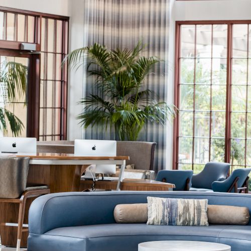 A stylish, sunlit lounge with blue sectional sofa, round ottoman, potted palm plants, and large windows framing a modern workspace.