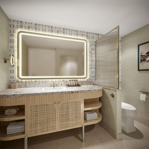 A modern bathroom with a wicker vanity, open shelves, a backlit rectangular mirror, patterned wallpaper, and a frosted glass door.