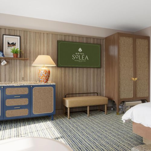 A hotel room with a blue dresser, wooden paneling, a bench, a wall-mounted TV showing &ldquo;SOLEA,&rdquo; and a bed at the right.