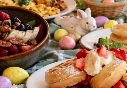 A festive Easter spread: berries, pancakes with cream and strawberries, pastel eggs, a bunny figurine, and waffles in a colorful brunch scene.