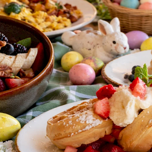 A festive Easter spread: berries, pancakes with cream and strawberries, pastel eggs, a bunny figurine, and waffles in a colorful brunch scene.