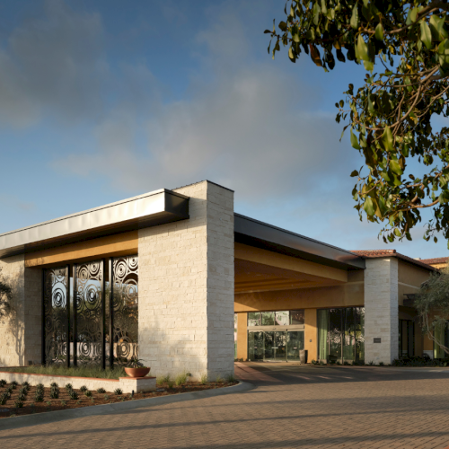 A modern low-rise building with large glass windows, stone walls, and a wide covered entrance surrounded by trees and a paved area.