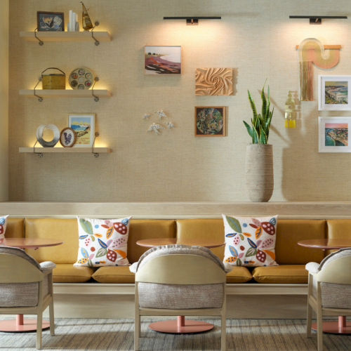 A modern, cozy lounge with a long beige sofa, colorful cushions, light wooden chairs, wall shelves, framed art, a tall plant, and ambient spotlights.