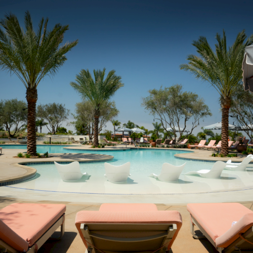 A resort-style pool area with palm trees, white lounge chairs, and a bright blue sky, perfect for sunbathing and relaxation.