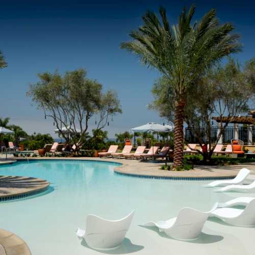 A luxury resort pool with clear water, white lounge chairs, palm trees, shaded seating areas, and sunny skies.