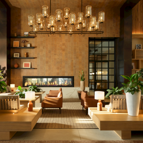 Cozy-modern lobby with warm lighting, a central fireplace, wooden benches, potted plants, and a grid of shelves along the back wall.