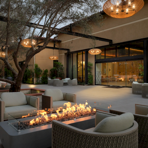 Cozy outdoor lounge with modern fire pits, wicker seating, soft lighting, and a shaded courtyard leading to an indoor seating area.