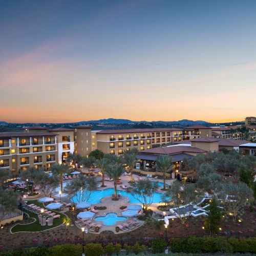 A resort-style pool area with lounge chairs, a central pool, palm trees, and surrounding low-rise buildings at sunset.
