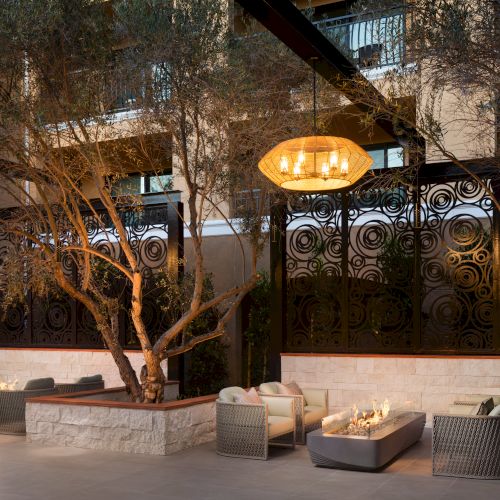 A stylish hotel courtyard with wicker lounge chairs, soft cushions, tree planters, and warm hanging lanterns overhead.