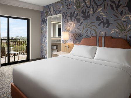 A bright hotel room with a large bed, patterned feature wall, a balcony door, and a small seating area outside.
