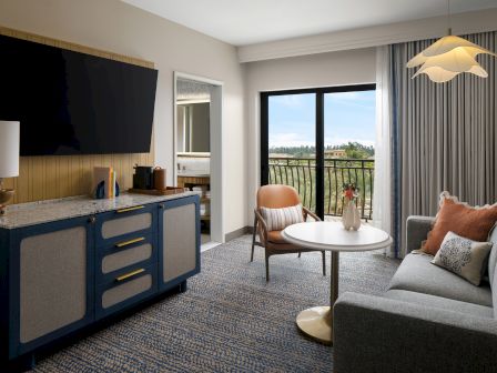 A modern hotel suite with a blue-white kitchenette, a flat-screen TV, a seating area, round table, and a balcony view through large windows.