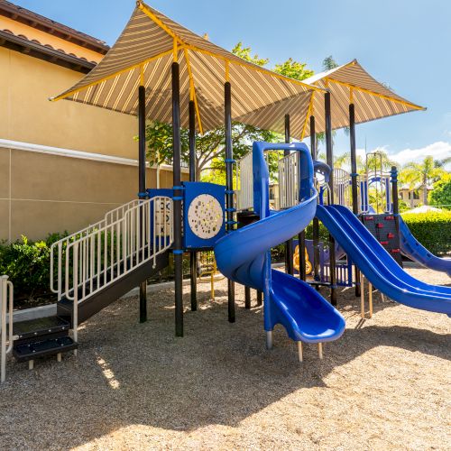 A blue multi-slide playground with shaded canopy, stairs, and climbing structures beside a tan building, on a sandy play area. ending now.