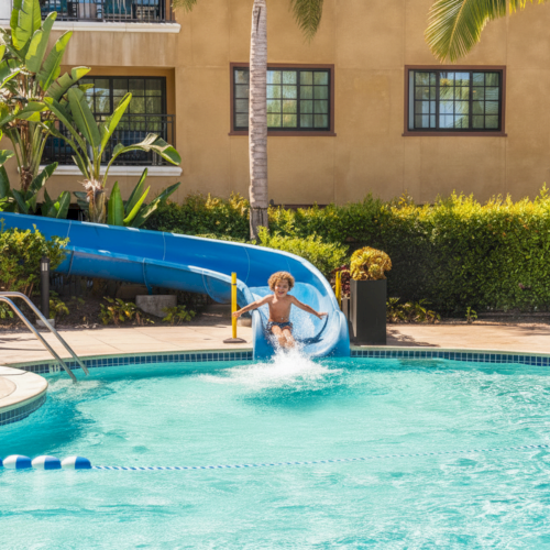 Two kids slide down a blue waterslide into a sunny hotel pool, palm trees and a tan building in the background.