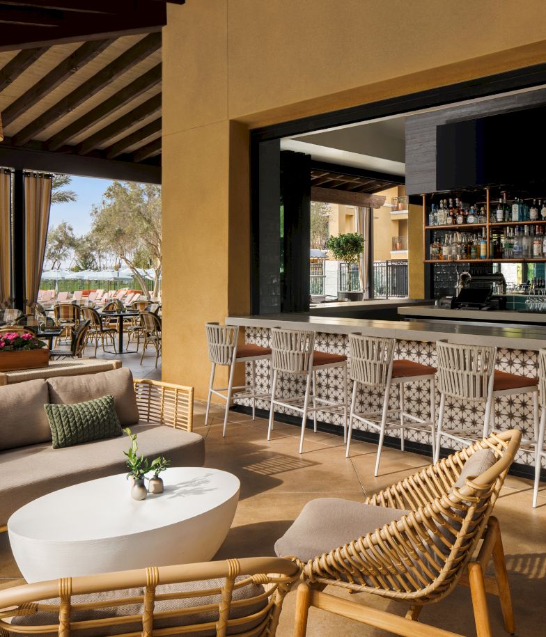 A stylish outdoor lounge and bar area with a seating sofa, wicker chairs, a white round coffee table, potted plants, and a long bar with white patterned stools under a shaded patio.