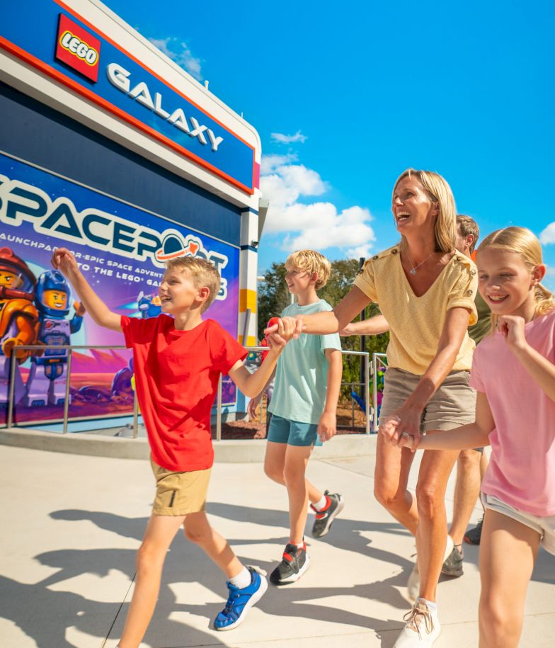 A family of four enjoys a sunny day at a LEGO-themed park, pointing excitedly toward Space-themed displays near a Galaxy storefront.