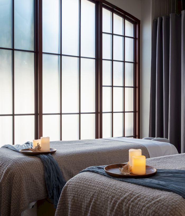 A serene spa room with two massage tables, candles, soft lighting, and a robe hanging by a windowed wall, creating a warm, relaxing vibe.