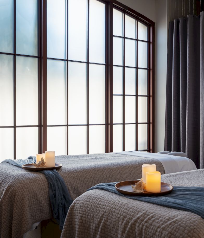 A serene spa room with two massage tables, candles, soft lighting, and a robe hanging by a windowed wall, creating a warm, relaxing vibe.