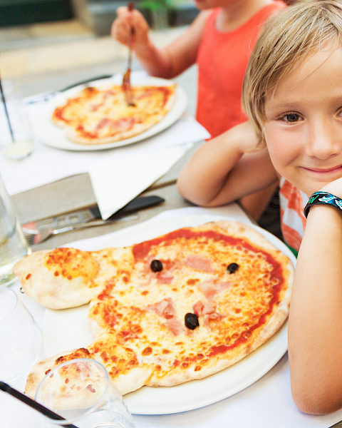 Three kids at a table with pizza, drinks, and a sunny outdoor setting; the girl in the foreground smiles with a watch on her wrist.