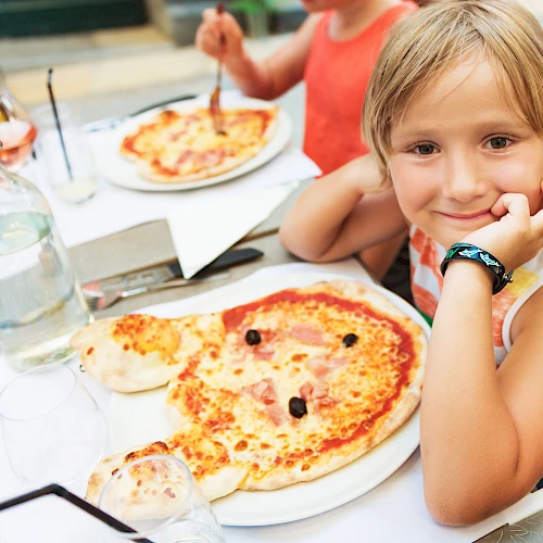 Three kids at a table with pizza, drinks, and a sunny outdoor setting; the girl in the foreground smiles with a watch on her wrist.