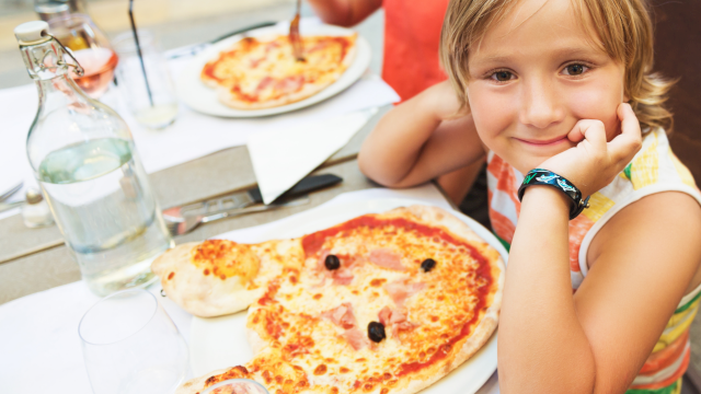 Three kids at a table with pizza, drinks, and a sunny outdoor setting; the girl in the foreground smiles with a watch on her wrist.