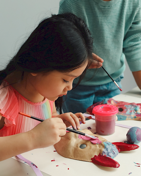A young girl paints clay with brushes at a table, with paints, tools, and a supervising adult nearby; creative art session in progress.