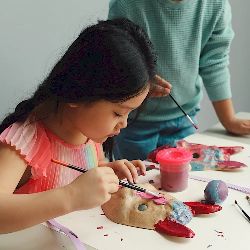A young girl paints clay with brushes at a table, with paints, tools, and a supervising adult nearby; creative art session in progress.