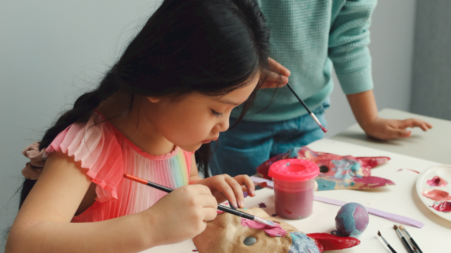 A young girl paints clay with brushes at a table, with paints, tools, and a supervising adult nearby; creative art session in progress.