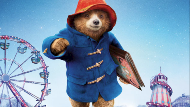 A sloth in a blue coat and red hat is holding a book, standing in a colorful amusement park with a ferris wheel and rides at night.