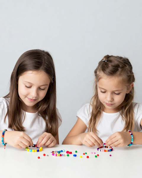 Two girls sit at a table stringing colorful beads to make bracelets while looking down at their craft supplies.
