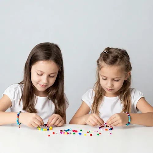 Two girls sit at a table stringing colorful beads to make bracelets while looking down at their craft supplies.