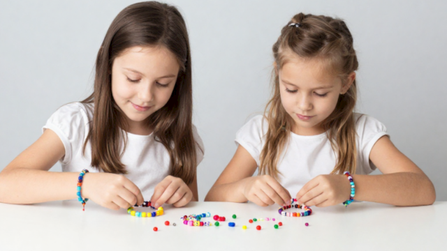Two girls sit at a table stringing colorful beads to make bracelets while looking down at their craft supplies.
