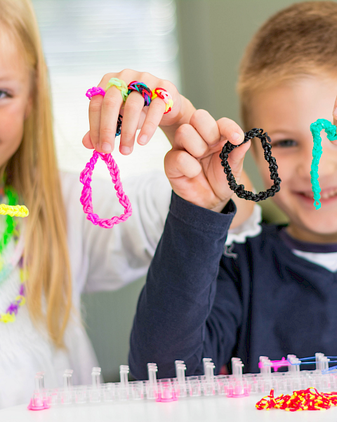 Two kids show off colorful bracelets they made with bright beads and strings, smiling proudly at their craft project.