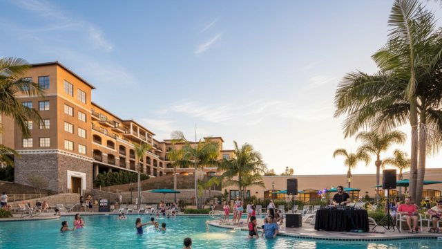 A sunny resort pool scene with people swimming and lounging, palm trees, and modern buildings in the background, bright and inviting.