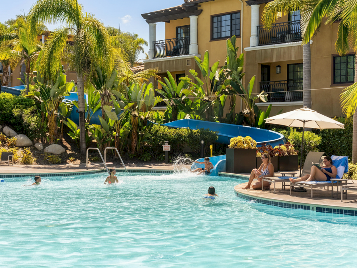 A sunny resort pool scene with palm trees, a water slide, lounge chairs, and people enjoying the refreshing day by the yellow building.