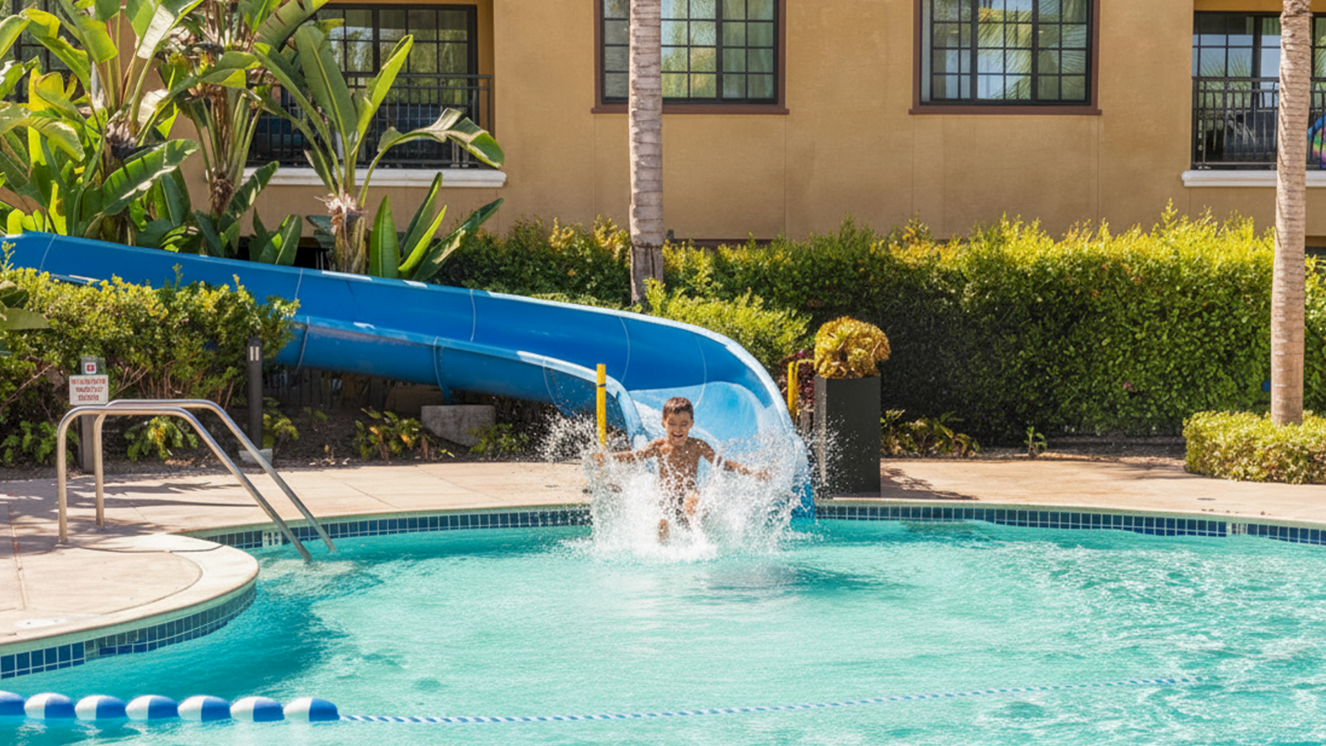A kid zooms down a blue water slide into a bright pool, splashing as he emerges near the edge.
