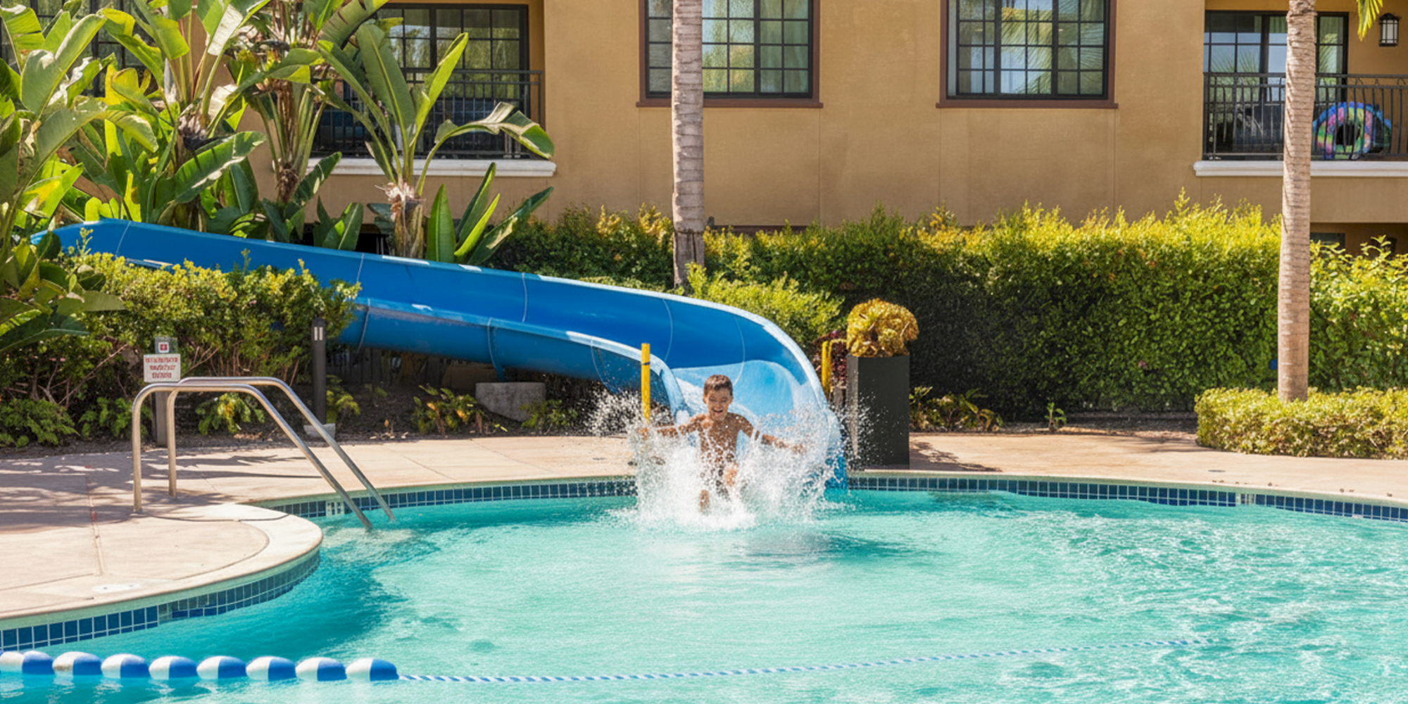 A kid zooms down a blue water slide into a bright pool, splashing as he emerges near the edge.