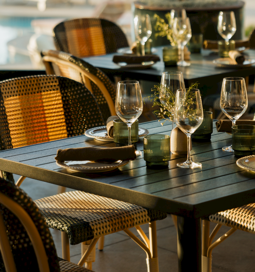Outdoor restaurant tables set with wine glasses, napkins, and cutlery, ready for guests on a sunny dining patio.