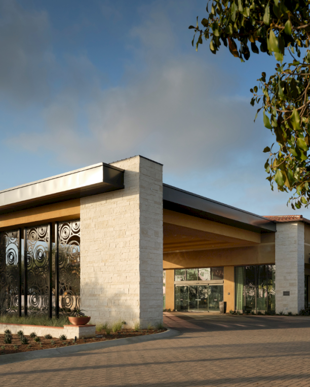 A modern low‑slung building with tall glass walls, stone columns, and a flat overhang sits in a sunlit, landscaped campus area.