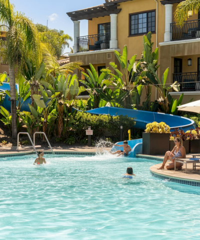 Poolside at a sunny resort with palm trees, people swimming, lounge chairs, and a yellow building in the background.