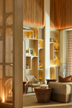 A stylish hotel lobby with warm wood tones, decorative partitions, built-in shelving, cozy seating, and a bright, airy atmosphere.