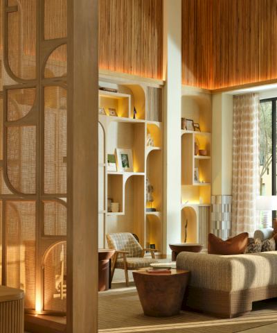 A stylish hotel lobby with warm wood tones, decorative partitions, built-in shelving, cozy seating, and a bright, airy atmosphere.