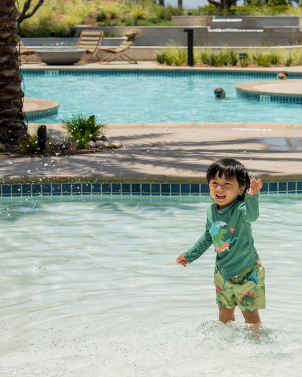 A little boy splashes happily in a sunny outdoor pool with lounge chairs and palm trees in the background, soaking up fun.