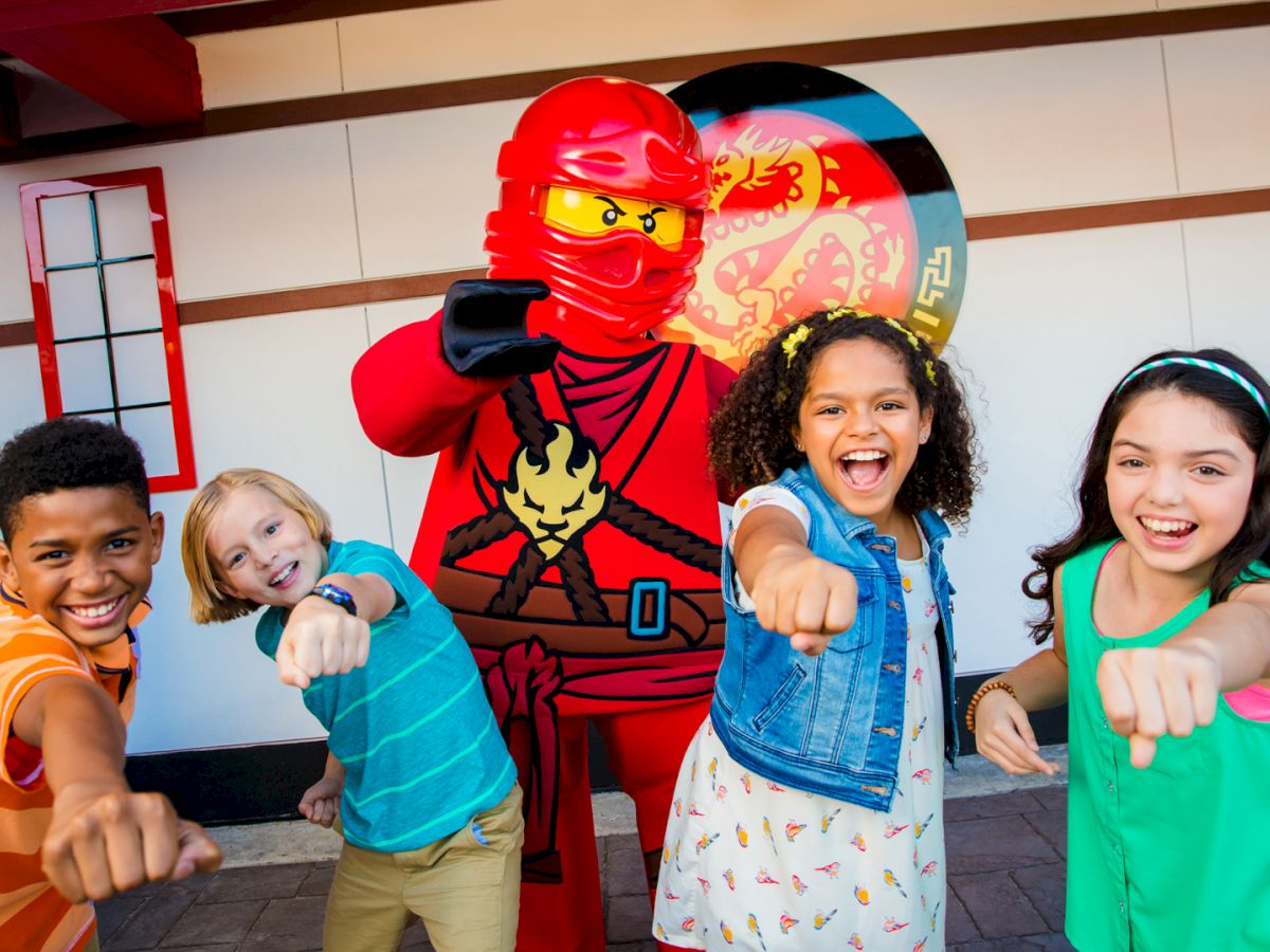 Kids laugh and point at the camera beside a red LEGO-style ninja character in a playful, colorful indoor scene.